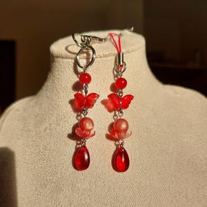 Op de afbeelding: Een paar rode oorhangers. Elke oorbel heeft een zilverkleurige metalen haak, een rode kraal, een rode vlinderbedel, een roze bloemvormige kraal en een druppelvormige rode kraal. De oorbellen worden gepresenteerd op een beige mannequinhoofd.