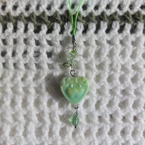 Puede incluir: Un dije en forma de corazón verde claro con un diseño de lazo, suspendido de un cordón verde. El dije está adornado con una pequeña hoja y cuentas de cristal. El fondo es un patrón de ganchillo blanco y verde.