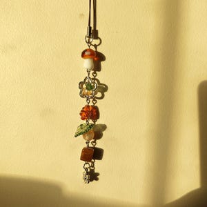 May include: A decorative charm with a brown cord, featuring a mushroom, flower, leaf, and square beads. The mushroom has a white stem and a brown cap. The flower has green and yellow accents. The leaf is green and orange.