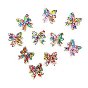 May include: Ten colorful confetti bow hair clips. The bows are made of clear resin with embedded confetti in shades of pink, blue, green, yellow, and orange. The bows are all different sizes and shapes.