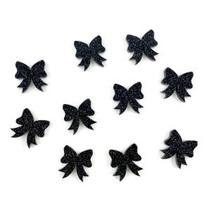 May include: Ten black glitter bow embellishments. The bows are all the same size and shape, and they are arranged in a random pattern on a white background.