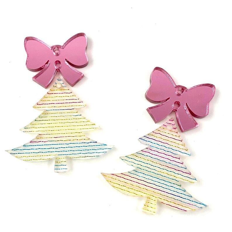 Puede incluir: Dos adornos de lazo rosa con purpurina con un dise&ntilde;o de &aacute;rbol de Navidad blanco con purpurina. Los &aacute;rboles tienen un patr&oacute;n de rayas multicolores.