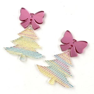 Puede incluir: Dos adornos de &aacute;rbol de Navidad de acr&iacute;lico con un lazo rosa en cada uno. Los &aacute;rboles est&aacute;n decorados con rayas arco&iacute;ris y purpurina.