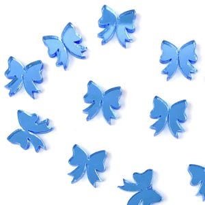 May include: A collection of light blue, bow-shaped acrylic embellishments. The bows are scattered across a white background, each with a reflective, mirrored surface. These decorative elements are ideal for crafting and adding a touch of elegance.