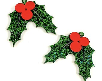 May include: Two Christmas holly leaf ornaments. Each ornament features a green glittery leaf design with red berries. The leaves have a jagged edge and a small hole at the top for hanging. The ornaments are set against a white background.
