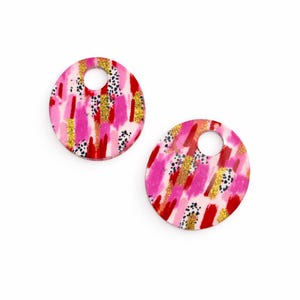 May include: Two round earrings with a hole at the top, featuring an abstract design. The earrings have pink, red, gold, and black brushstrokes. They are set against a white background.