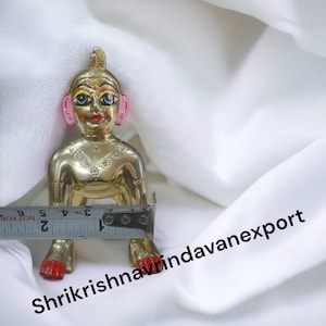 May include: A small, gold-colored statue of a baby Krishna, sitting with legs crossed. The statue has painted details, including blue eyes, pink ears, and red feet. The statue is approximately 4 inches tall.