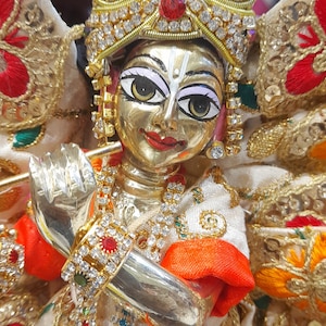 May include: A gold deity statue, wearing a jeweled crown and elaborate garments. The statue has a calm expression and is embellished with colourful gemstones and gold detailing. The figure's hands are in a prayer position.