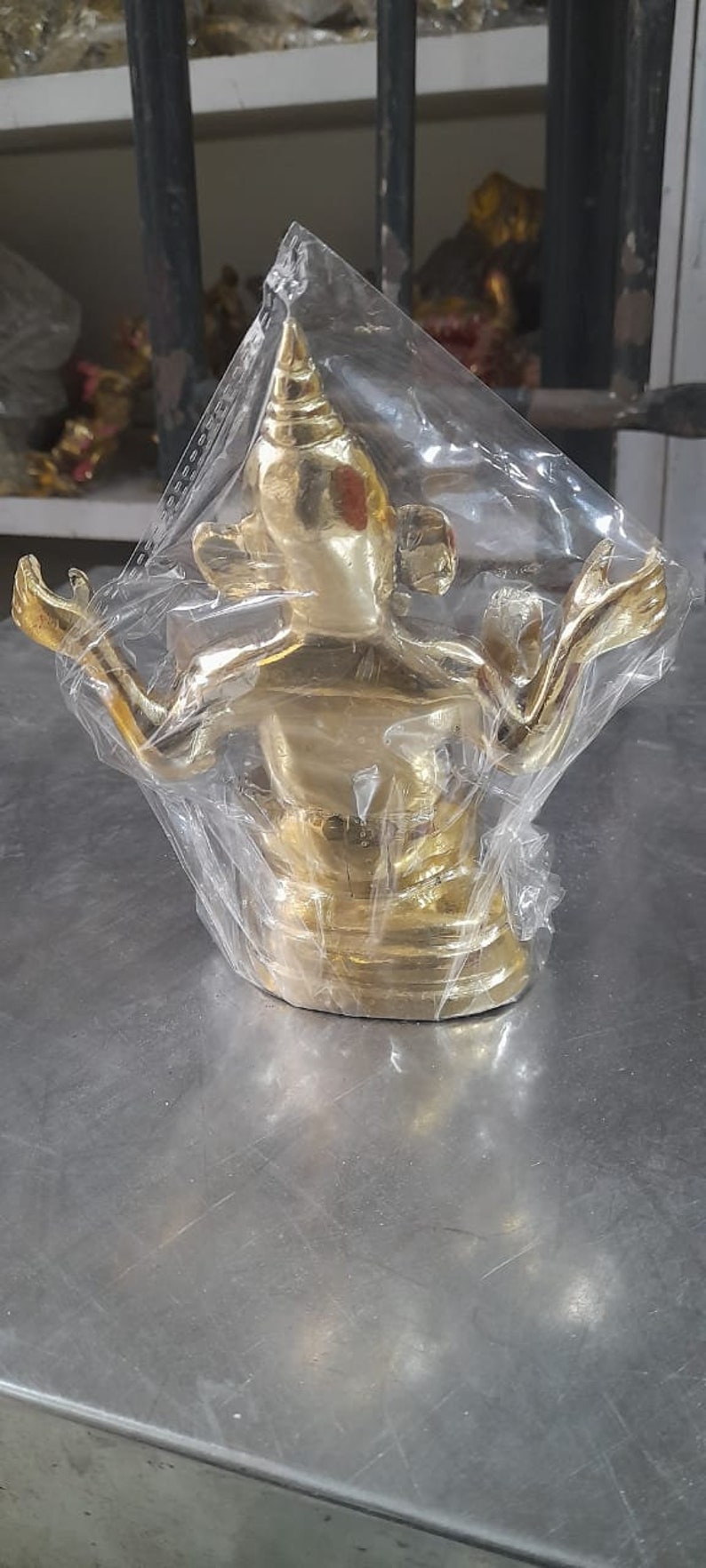 May include: A gold-colored metal statue of a deity with a pointed head and outstretched arms. The statue is wrapped in clear plastic.