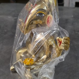 May include: A gold-colored Ganesha statue with red and yellow paint details, wrapped in clear plastic.