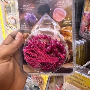 May include: A clear plastic blister pack containing a strand of dark pink beads with a matching pink tassel. The packaging features printed text and images of various crystals, including amethyst, black obsidian, rose quartz, and citrine.