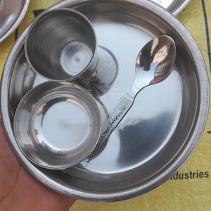 May include: A set of stainless steel Indian dining utensils. The set includes plates, small bowls, a cup, and spoons. The utensils are arranged on a yellow surface. The spoons have decorative handles.