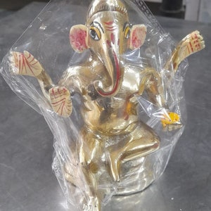 May include: A gold-colored Ganesha statue with red and yellow accents. The statue is sitting on a base and has its arms raised. The statue is wrapped in clear plastic.