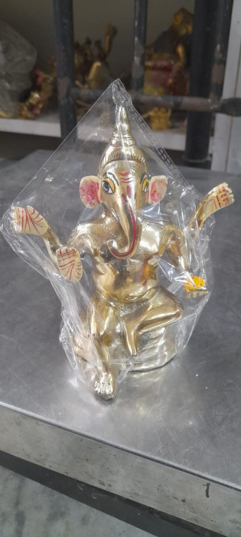 May include: A gold-colored Ganesha statue with red and yellow accents. The statue is sitting on a base and has its arms raised. The statue is wrapped in clear plastic.