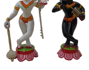 May include: Two painted figurines of Hindu deities, one white and one black, standing on lotus-shaped bases. The white figurine holds a staff with a pearl at the top, and the black figurine holds a flute. Both figurines are decorated with colourful details.