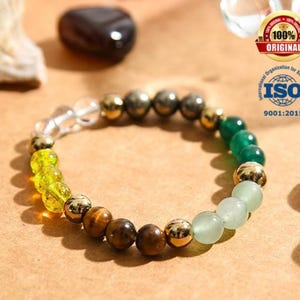 Könnte beinhalten: Ein Perlenarmband mit einer Vielzahl von Edelsteinen und goldfarbenen Perlen. Das Armband enthält klare, gelbe, braune, grüne und hellgrüne Perlen. Das Armband wird auf einer hellbraunen Oberfläche präsentiert.