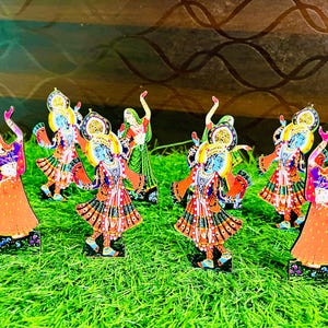 May include: A collection of colorful, decorative figurines depicting traditional Indian dancers. The figurines are on a green surface, with vibrant orange, blue, and green clothing. The figures have detailed facial features and ornate headdresses.