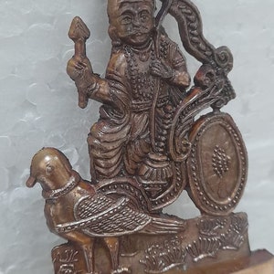 Könnte beinhalten: Eine kleine, bronzefarbene Statue einer Gottheit, die auf einem Vogel reitet. Die Gottheit trägt eine Krone und hält einen Bogen und Pfeil. Die Statue steht auf einem Sockel mit der Inschrift "Om Jay Shani Dev Om".