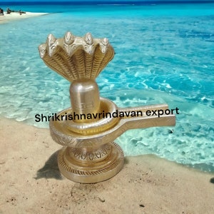 Könnte beinhalten: Eine goldfarbene, verzierte Shiva Lingam-Statue mit einer fünfköpfigen Kobrahaube, die auf einem Sandstrand mit blauem Wasser im Hintergrund ruht. Die Statue ist mit aufwendigen Schnitzereien verziert und hat einen Sockel mit einem Griff. Der Text "Shrikrishna Vrindavan Export" ist auf der Statue sichtbar.