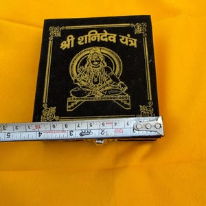 May include: A square black box with gold-coloured printed text and a central deity illustration. A metal measuring tape is positioned across the front, showing a width of approximately 10 cm. The box features ornate corner details.