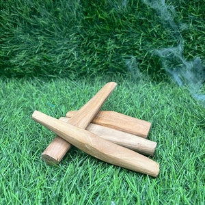 May include: A collection of light brown wooden incense sticks, some with pointed ends, arranged on a bed of green artificial grass. Wisps of smoke are visible in the background, suggesting the sticks are burning.