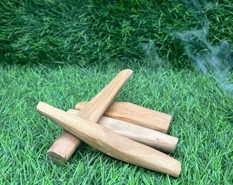Può includere: Una collezione di bastoncini di incenso in legno marrone chiaro, alcuni con estremità appuntite, disposti su un letto di erba artificiale verde. Volute di fumo sono visibili sullo sfondo, a suggerire che i bastoncini stiano bruciando.