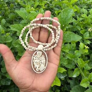 Puede incluir: Un collar con un colgante redondo de madera clara que presenta una ilustración grabada en marrón oscuro de dos figuras de pie una al lado de la otra. El collar está compuesto por pequeñas cuentas redondas de color blanco roto.