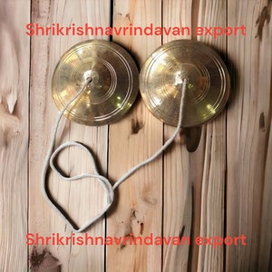 May include: Two gold-colored cymbals with a white string connecting them. The cymbals are on a wooden surface. The text "Shrikrishna Vrindavan Export" is visible at the top and bottom of the image.