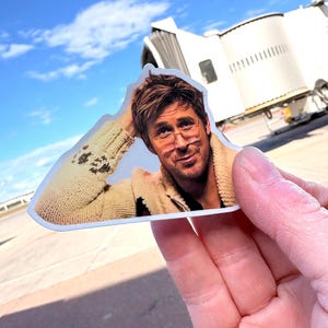 May include: A die-cut sticker featuring a man with glasses wearing a beige sweater. The sticker is held up against a backdrop of a blue sky and an airport terminal.