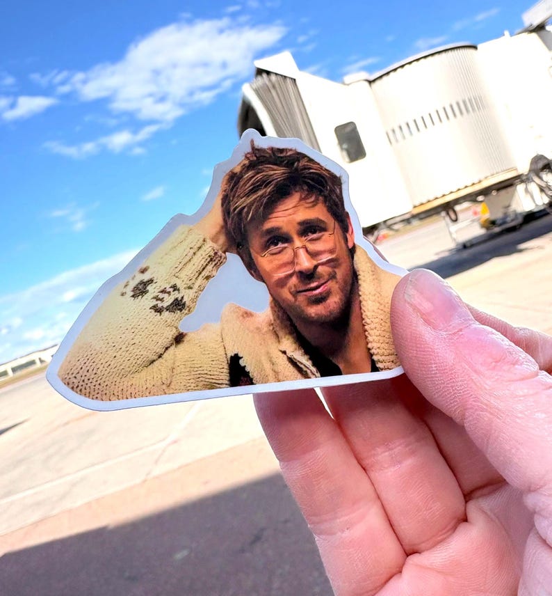 May include: A die-cut sticker featuring a man with glasses wearing a beige sweater. The sticker is held up against a backdrop of a blue sky and an airport terminal.