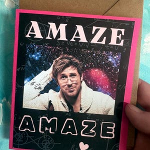 May include: A rectangular greeting card with a black background featuring white mathematical formulas and a central photograph of a person with brown hair wearing a cream-colored sweater against a galaxy backdrop. The word AMAZE is printed in pink.