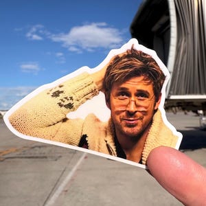 May include: A die-cut sticker featuring a man with glasses, wearing a beige sweater. The sticker is held up against a blue sky and airport background. The man is smiling and has his hand in his hair.