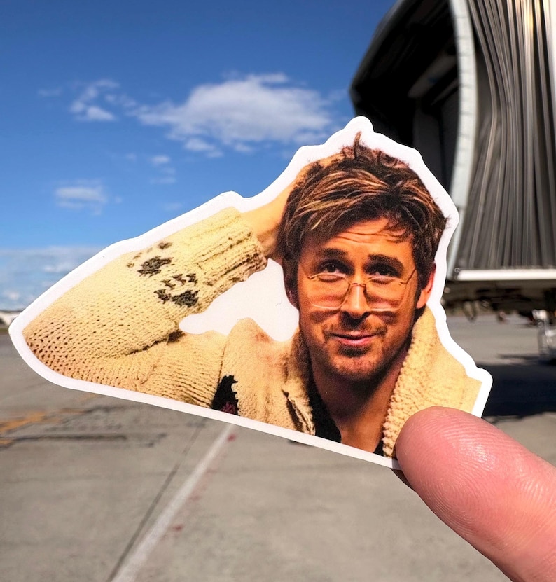 May include: A die-cut sticker featuring a man with glasses, wearing a beige sweater. The sticker is held up against a blue sky and airport background. The man is smiling and has his hand in his hair.
