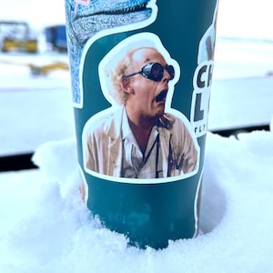 Puede incluir: Una taza de viaje de color turquesa con una pegatina de Doc Brown de Regreso al futuro. La pegatina muestra a Doc Brown con gafas protectoras y una bata de laboratorio blanca. El texto "FLY" es visible en la pegatina.