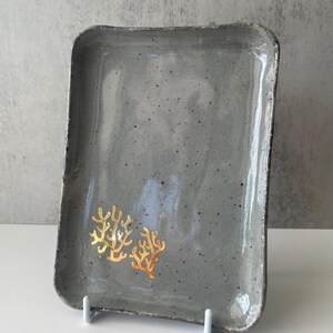 May include: A rectangular ceramic serving tray in a muted grey with a speckled finish. Two gold coral designs are featured on the tray's surface. The tray is supported by a white stand, showcasing a handcrafted aesthetic.