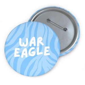 May include: A round pin-back button featuring a light blue and white wavy striped pattern. The text WAR EAGLE is printed in bold white letters across the center. The back of the button is metallic silver with a standard pin closure mechanism.
