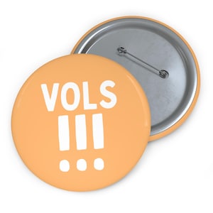 May include: A round pinback button with a light orange background featuring the white text VOLS followed by three vertical bars and three dots arranged in a horizontal row. The back of the button is silver metal with a pin mechanism attached.