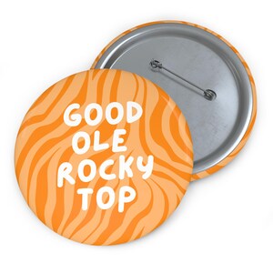 May include: A round pin-back button featuring an orange and light orange wavy striped pattern. The text GOOD OLE ROCKY TOP is printed in white, rounded, sans-serif capital letters across the center of the button face. The back shows a metal pin.
