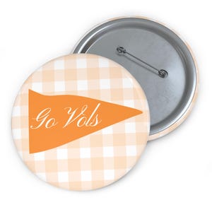 Puede incluir: Una chapa redonda con un patrón de cuadros vichy en color naranja claro y blanco. En el centro, un gráfico de banderín naranja lleva el texto blanco Go Vols en letra cursiva. La parte posterior muestra un mecanismo de alfiler metálico.