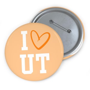 May include: A round pin-back button with a light orange background. The front features the white text I and UT in a block font, separated by an orange hand-drawn heart symbol. The back of the button is silver metal with a horizontal pin mechanism.