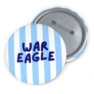 May include: A round pin-back button featuring vertical light blue and white stripes. The text "WAR EAGLE" is printed in dark blue, rounded, sans-serif capital letters across the center. The back of the button shows a silver metal pin mechanism.