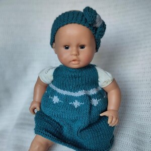 Peut inclure: Une robe en tricot bleu sarcelle avec des accents blancs et un bandeau assorti en bleu sarcelle avec un nœud gris. La robe est portée par une poupée aux yeux bruns et au teint clair.