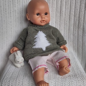 Könnte beinhalten: Eine Puppe trägt einen grünen Strickpullover mit einem weißen Weihnachtsbaum-Motiv und eine rosa Hose. Die Puppe hält ein weißes Stoffkaninchen.