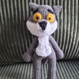 May include: A crocheted wolf toy in grey and white, with yellow eyes and a black nose. The wolf has a white chest and is standing upright. The background is a green corduroy fabric.