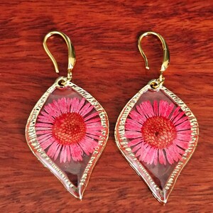 May include: A pair of gold-coloured dangle earrings. Each earring features a pink flower encased in clear resin within a teardrop-shaped gold frame. The earrings have a hook closure.