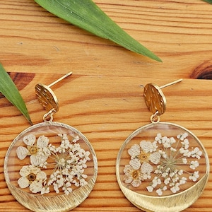 May include: A pair of gold-coloured earrings with a circular design. Each earring features a clear resin circle with dried white flowers and a textured gold-coloured metal frame. The earrings have a post-back closure.