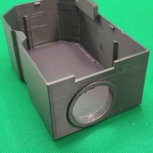 Pode incluir: Um objeto retangular cinza escuro impresso em 3D com uma parte superior aberta e uma abertura circular em um lado. O objeto tem formato de caixa com vários suportes estruturais internos e uma superfície texturizada.