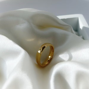 Puede incluir: Un anillo dorado con una superficie lisa y pulida. El anillo se encuentra sobre un fondo de tela de satén blanco, creando un contraste de texturas. El diseño del anillo es simple y elegante.