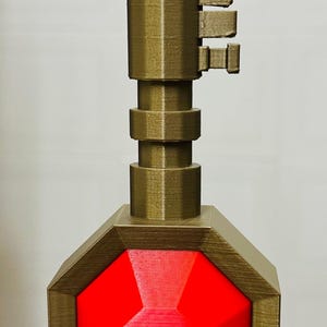 May include: A gold-colored key with a red, octagonal gem at the end. The key has a pointed top and a series of ridges along the shaft.