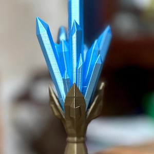 May include: A 3D printed crystal cluster with a gold base. The crystals are a light blue color and are arranged in a cluster on top of the base.